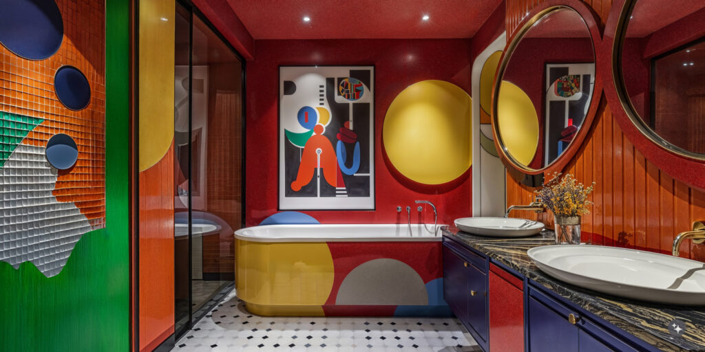 Pop art bathroom with black and white pattern floor tiles, bold geometric wallcovering in vibrant shades of green,orange, yellow, red and blue. A shower, a large painted tub and double sink vanity. 