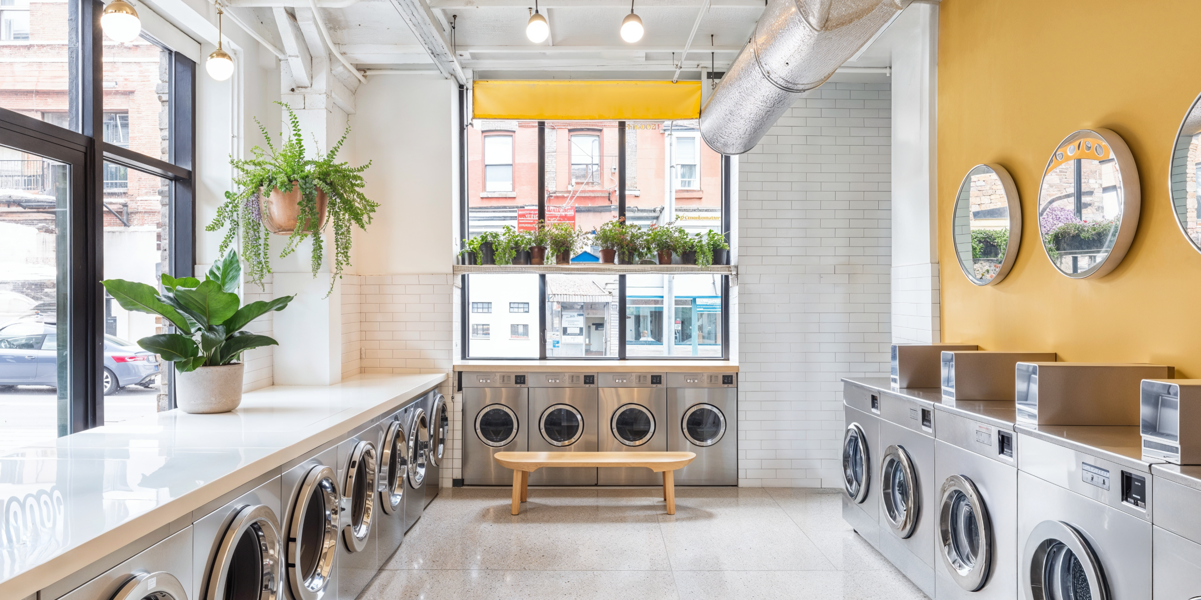 Modern laundromat with natural lighting, big windows, white tile floors, contemporary washers and dryers, tiled walls, bright accent wall, pendant light, and exposed ceilings.