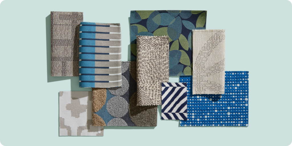 A photograph of a flatlay of a variety of different material samples in various colors, shapes and textures to represent the Material Bank Curated Collection Bold Impressions.It features a light blue background with samples in blue, gray, green, beige and white tones.