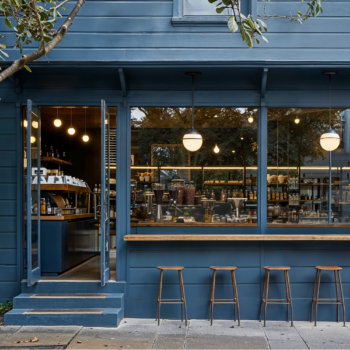 Modern Industrial Coffee Shop exterior painted in a deep blue, with wood stools and wood bar on the exterior. through the windows you can see racks of supplies and pendant hanging lights.