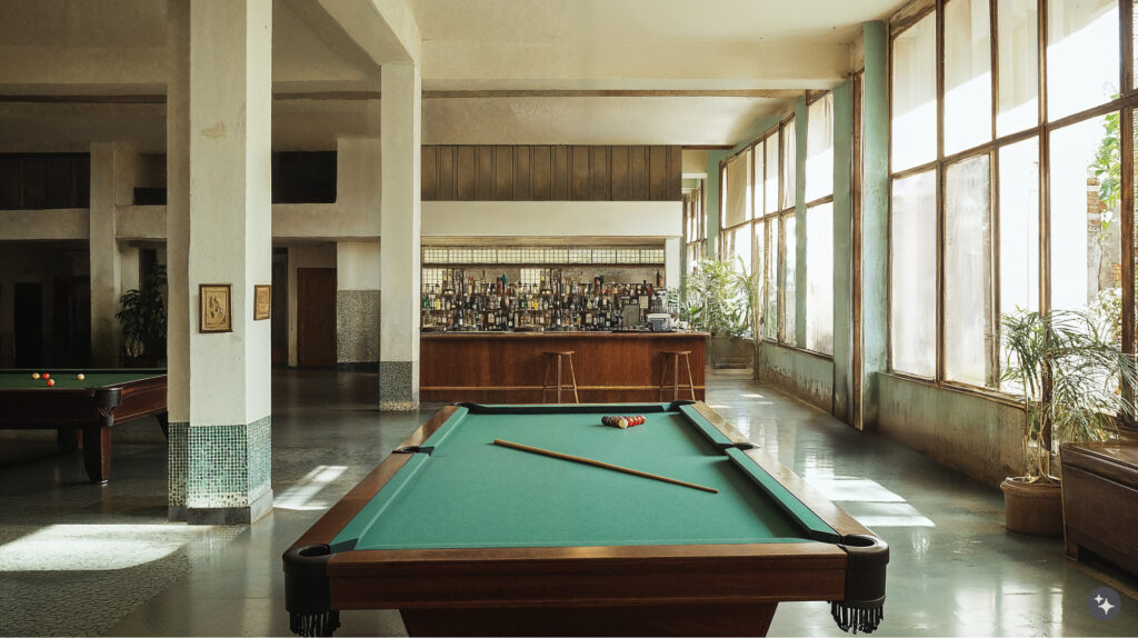 Cuban Design pool hall. A pool table sits directly in hte middle of the image and you can see additional pool table in the background along with a bar made of wood paneling. Large windows letting in light and mosaic tile detailing throughout the room. 