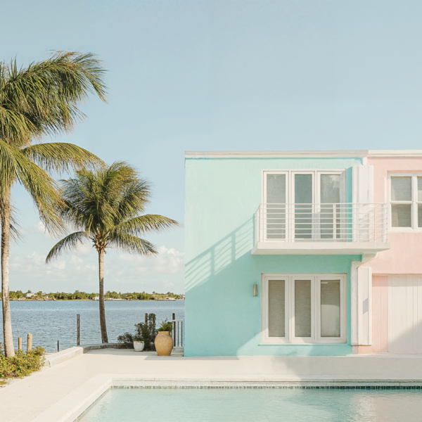 Key West design of a residential apartment building. Pastel blue and pink paint on the exterior of the building surrounded by the ocean in the background and in the foreground is a pool surrounded by lounge chairs and palm trees.