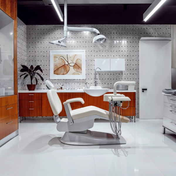 Mid-century modern dental offices with a contemporary dental chair, overhead lighting, sterile white tile flooring, wood cabinetry balanced with white cabinetry, and subtle black and white patterned wallpaper.