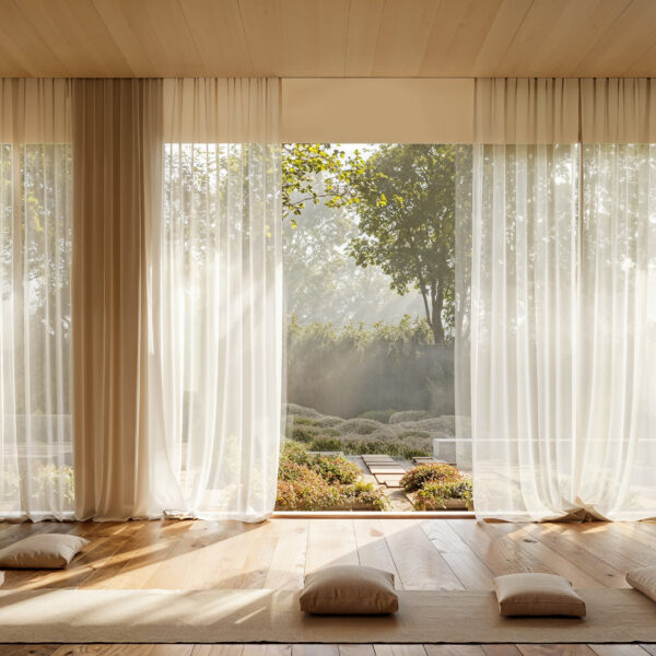 Meditation room with wood floors, woven mats and neutral colored pillows for sitting are place on the floor. A floor to ceiling wall of windows runs along one side of the room and sheer off white curtains frame sections of the glass which is looking out over a zen garden with stone pathways, plant life and a water feature.