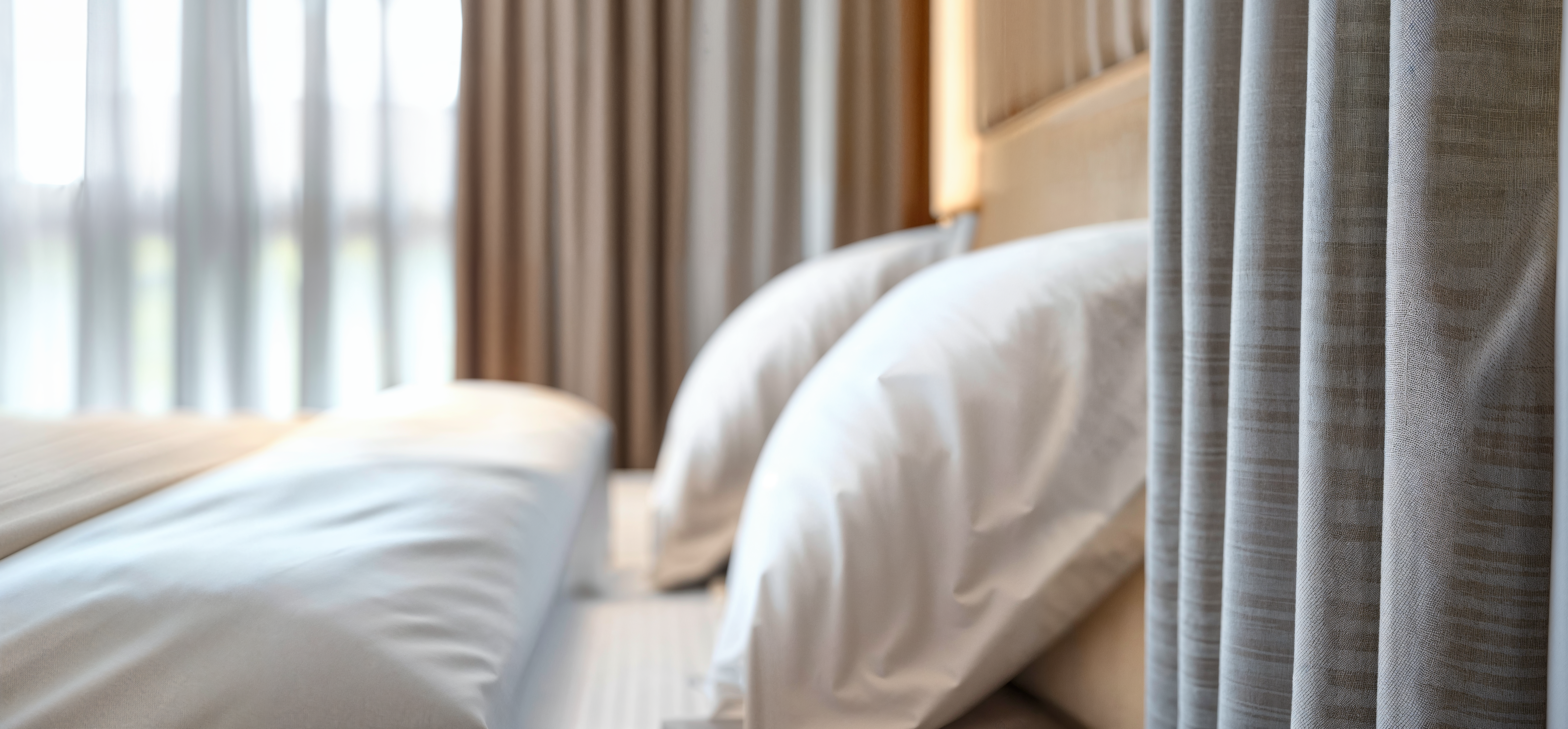 A close up photograph of a hotel bed slightly out of focusing highlighting the white and tan antimicrobial textiles. A large window covered with sheer curtains makes the room feel bright and airy.