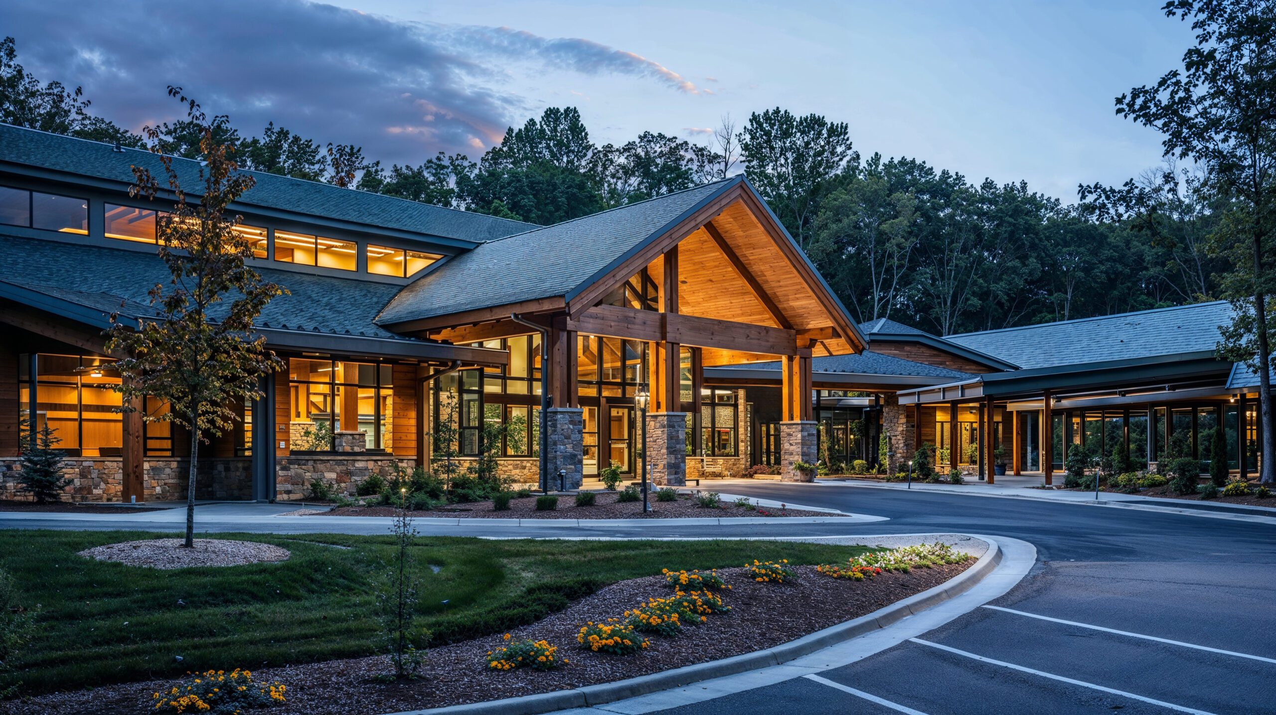 A modern, senior living facility with an exterior that features large windows and wooden accents, nestled in the woods. The entrance is decorated with beautiful flowers and plants, creating a welcoming atmosphere as you enter through its grand entryway and open parking lot.