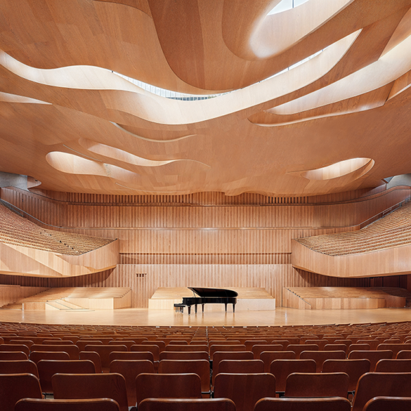 An interior of a concert hall featuring an open air style with a large space, a wooden ceiling adorned by undulating curves that resemble waves used as an innovation in architectural sounds design and seats arranged in rows. In front is a grand piano placed on stage.