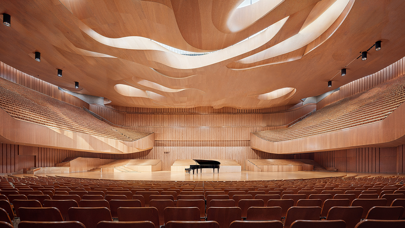 An interior of a concert hall featuring an open air style with a large space, a wooden ceiling adorned by undulating curves that resemble waves used as an innovation in architectural sounds design and seats arranged in rows. In front is a grand piano placed on stage.