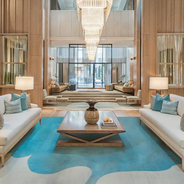 An example of a lobby as a living room with beige couches with wood coffee table with a plush beige and blue rug matching the blue accent pillows lining the couch creating a living room environment in a hotel lobby.