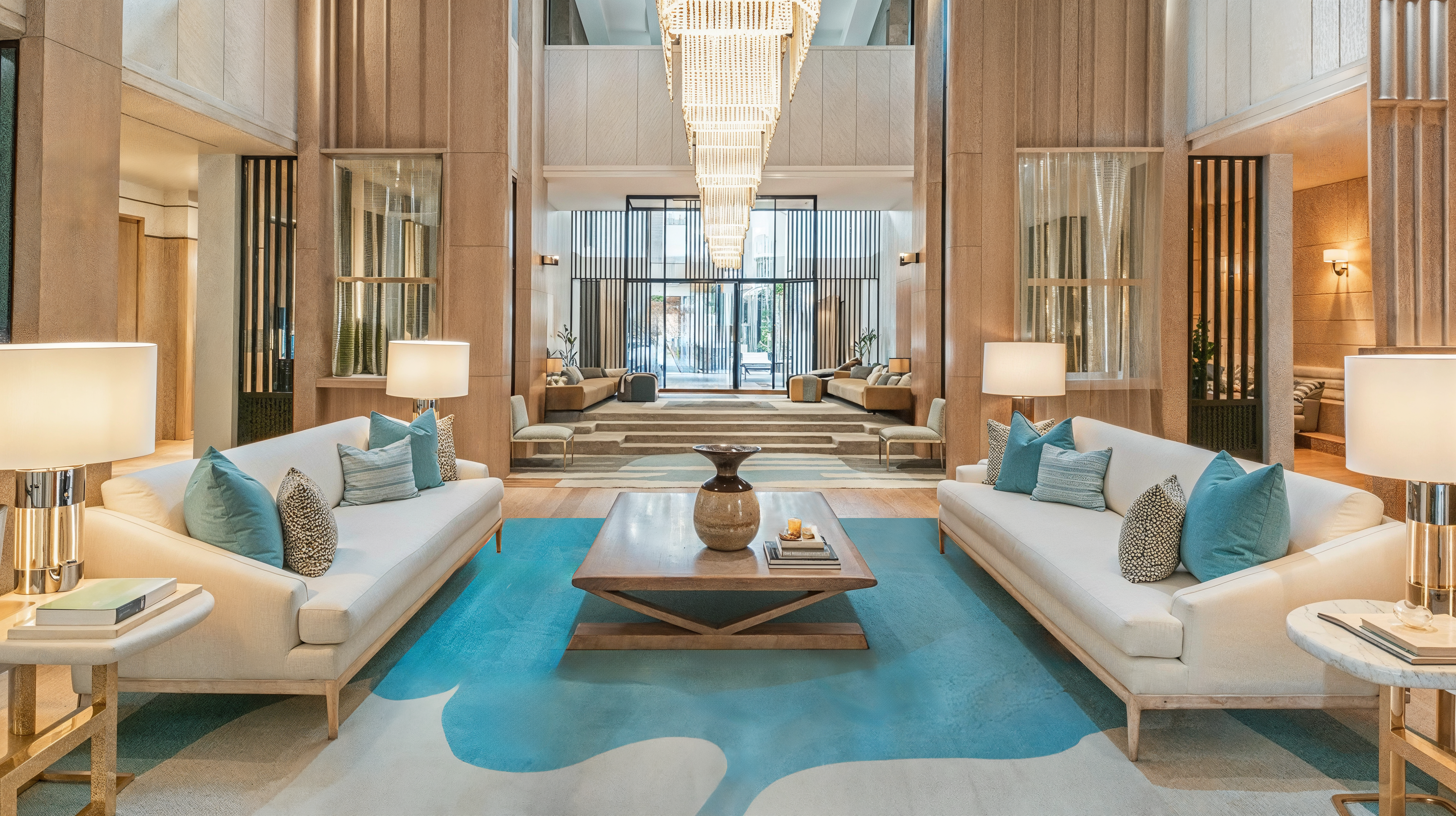 An example of a lobby as a living room with beige couches with wood coffee table with a plush beige and blue rug matching the blue accent pillows lining the couch creating a living room environment in a hotel lobby.