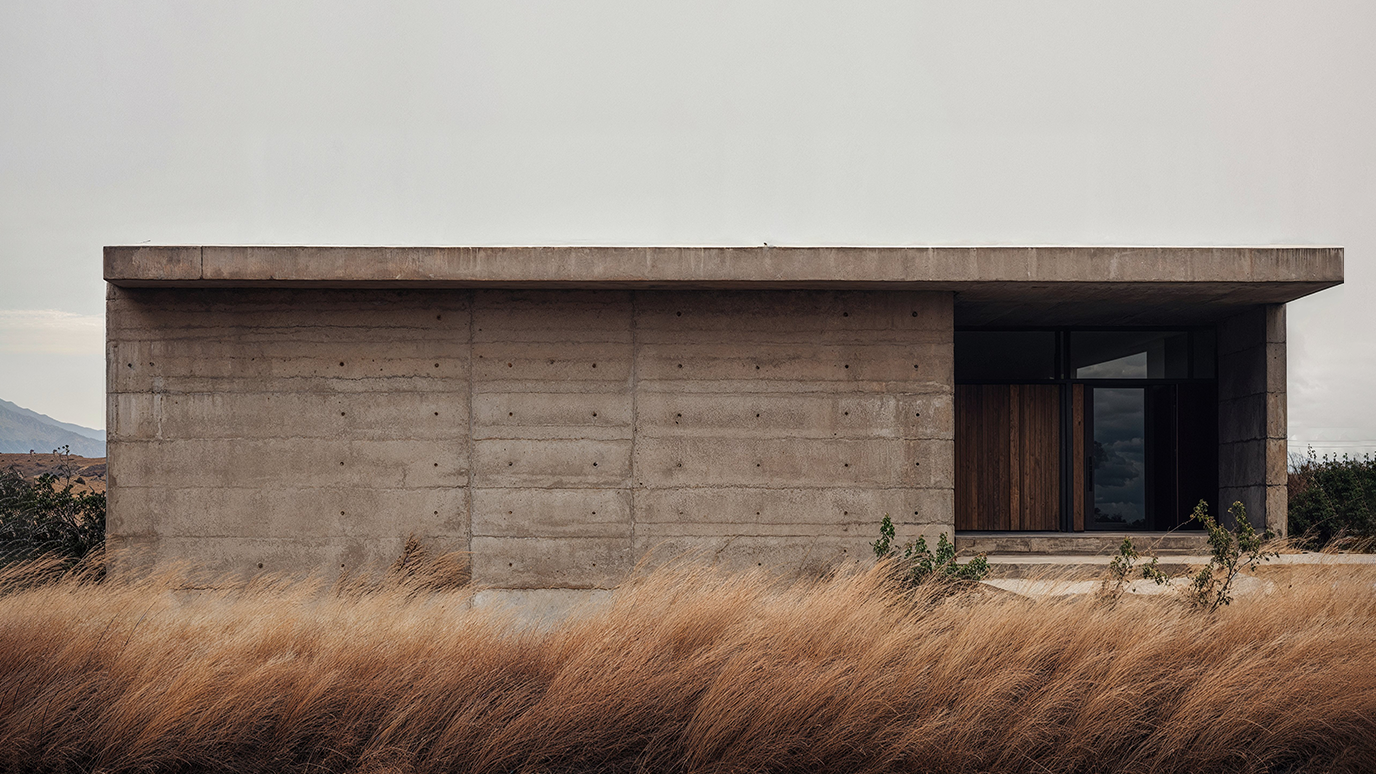 A photograph of a small, simple one-story concrete house, large windows, and a door on the right side, surrounded by tall grasses.