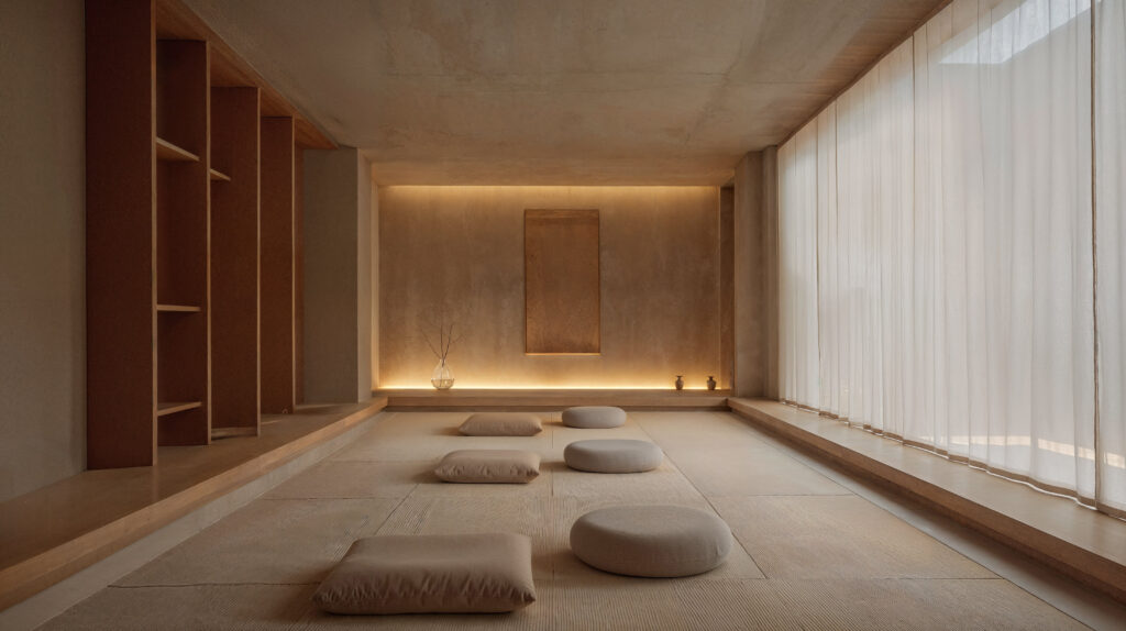 A room for meditation designed in the style of Japandi including beige and neutral colors, natural wood accents with woven rugs on the floor and neutral colored pillows for sitting. On one side of the room are floor to ceiling windows covering one wall and the windows are covered in  sheer curtains. 