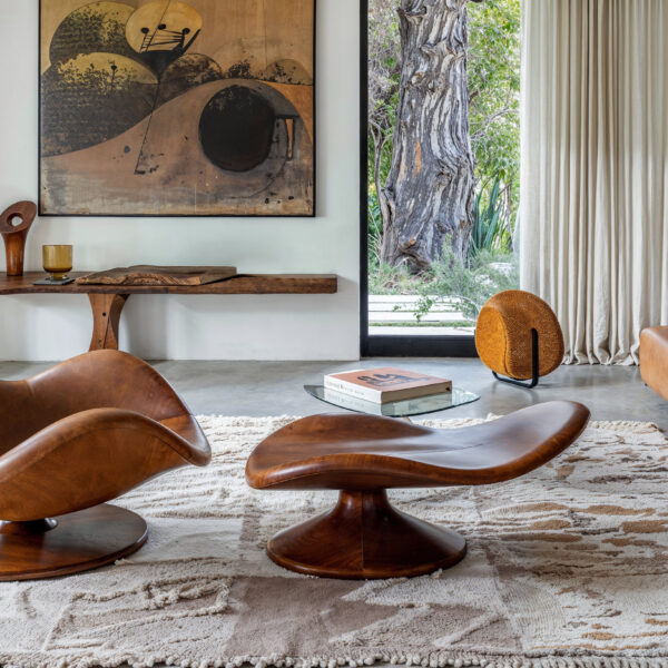 A modern lounge chair in brown leather sitting in the middle of the room atop a white and beige rug over concrete floors. A brown leather sofa sits against a wall with large floor to ceiling windows creating a bright, natural space.