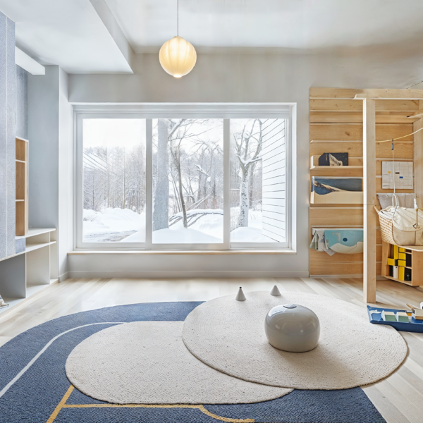 A posh playroom with light wood flooring, with several different shaped area rugs laying over it. Wood paneled walls on one side of the room that have built in shelving and racks and soft blue wallcovering with a delicate pattern on the other side of the room. Large windows looking outside.