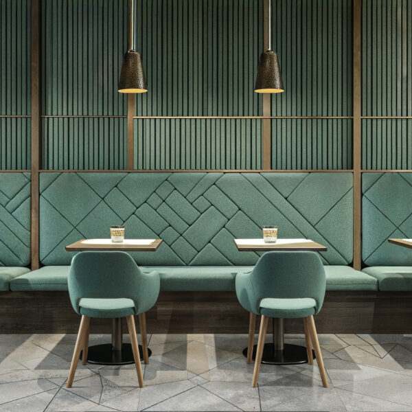 A restaurant with unique sound absorbing materials. Muted teal wall paneling and upholsetery on four 2 top tables with single chairs on one side and booth seating running along the back wall on the other.