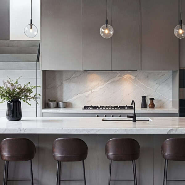 A minimalist kitchen with grey cabinets, white marble countertop and dark brown leather barstools, black pendant lights above kitchen, stairway on left side leading upstairs.