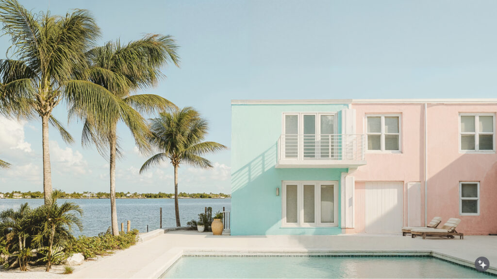 Key West design of a residential apartment building. Pastel blue and pink paint on the exterior of the building surrounded by the ocean in the background and in the foreground is a pool surrounded by lounge chairs and palm trees.