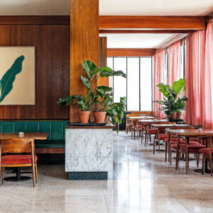 A Brazilian Modernist Designed restaurant with stone tile, wood tables and chairs, wood wall panels, green leather upholstery and foliage surrounding.