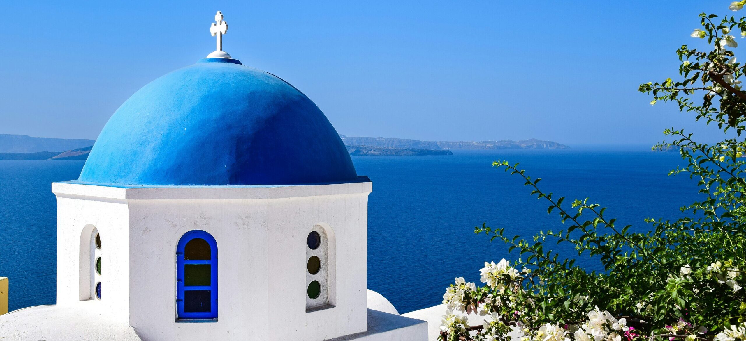 A photograph of Santorini Design featuring a domed building with a vibrant blue roof and door and crisp while building. Overlooking the sea.