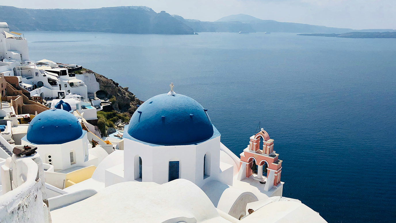 Photograph of Santorini, Greece with white buildings and blue domes overlooking the sea, with small church in foreground