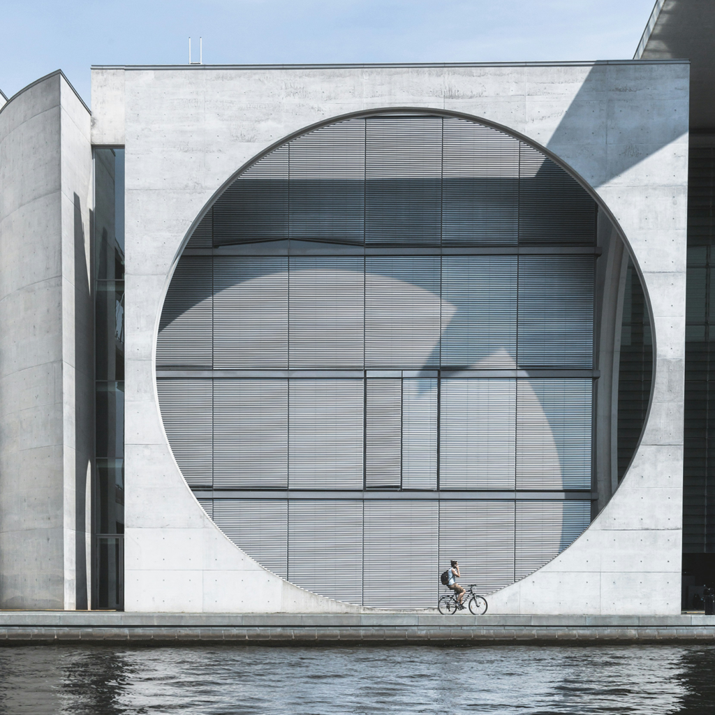 A modern designed concrete building with a large round window. A man riding a bicycle along the waterfront running in front of the building. 