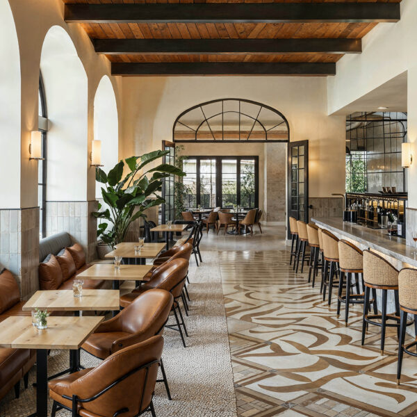 A restaurant showcase tile and stone in hospitality design with beige and brown abstract pattern tile on part of the floor and small beige mosaic stone on another. A gray natural stone countertop at the bar with small tables sitting across from it.