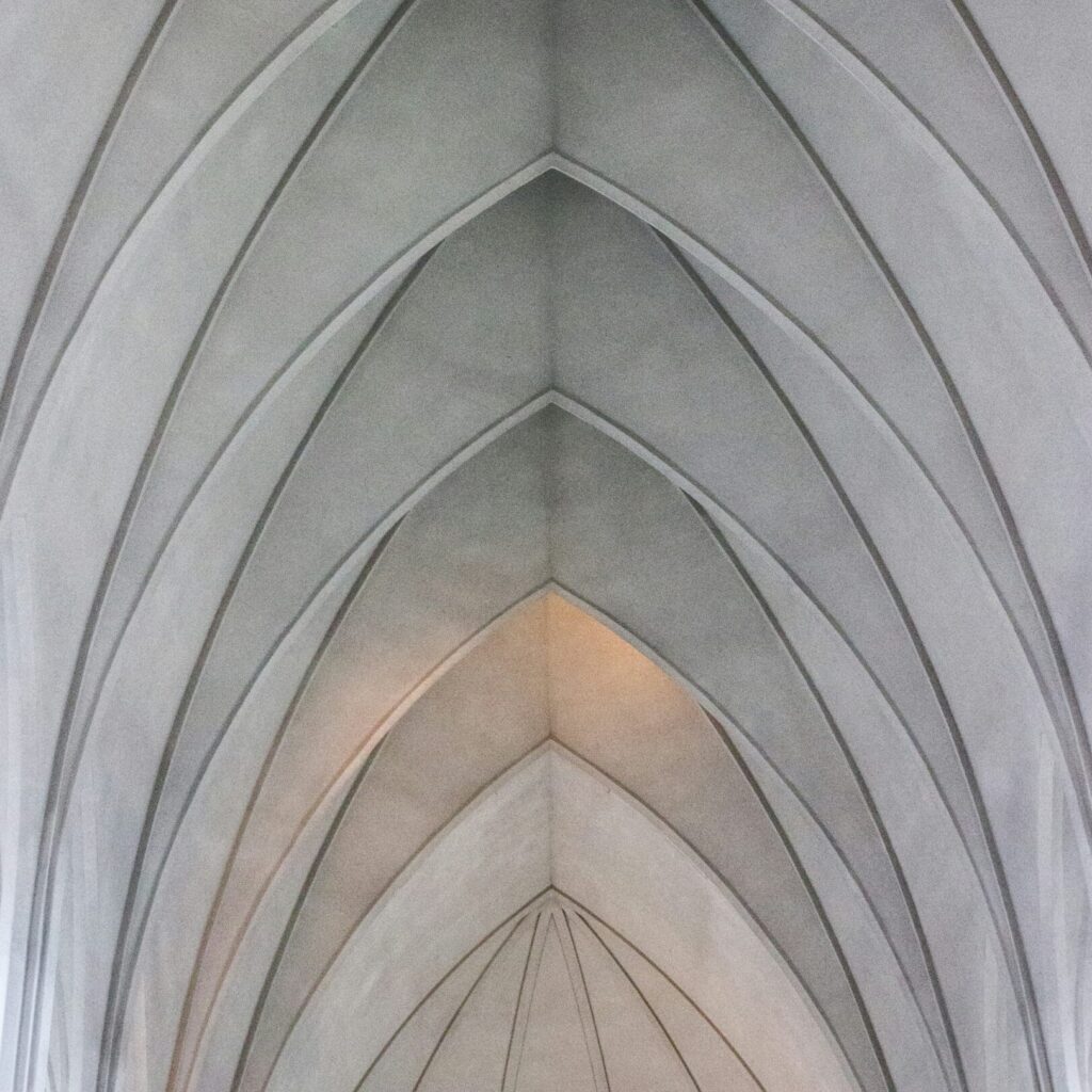 A photograph of the ceiling in Hallgrímskirkja, a cathedral in Reykjavík, Iceland. The arches form patterns that resemble simple triangles and can be seen from below. Soft lighting illuminates the grey concrete walls with subtle light reflections.