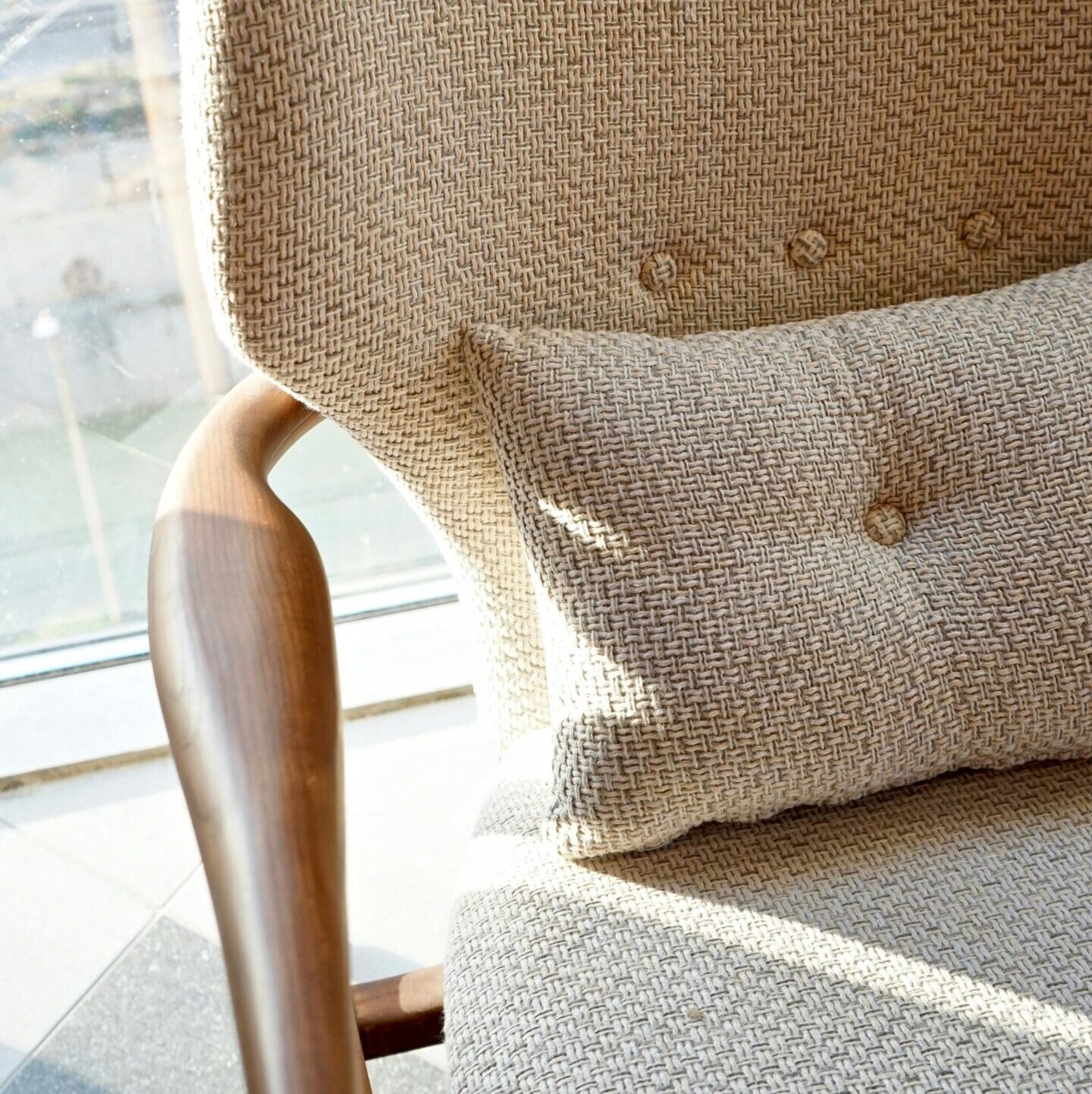 Close up of the armrests and cushion on an elegant, wooden chair with linen fabric in a light beige color, with buttons that add texture to its design. The background is a bright window casting gentle shadows across the interior of the room. This scene highlights how the soft fabric perfectly blends into the natural wood material, creating a harmonious atmosphere.