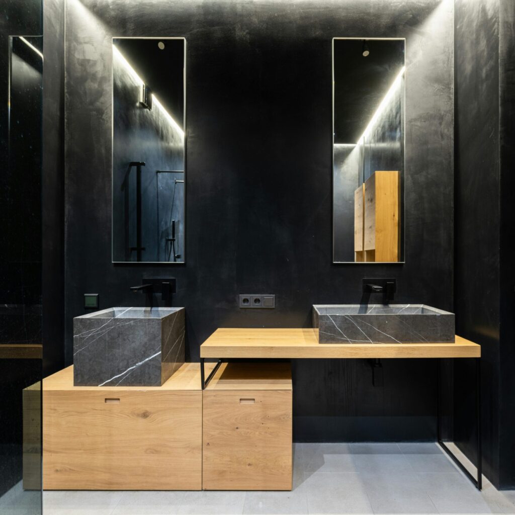 Modern interior design of the bathroom in black and oak wood, with two rectangular sculpture sinks in dark grey marble, wall mirror above it, dark gray walls, small wooden storage cabinet under the counter top, ambient lighting.