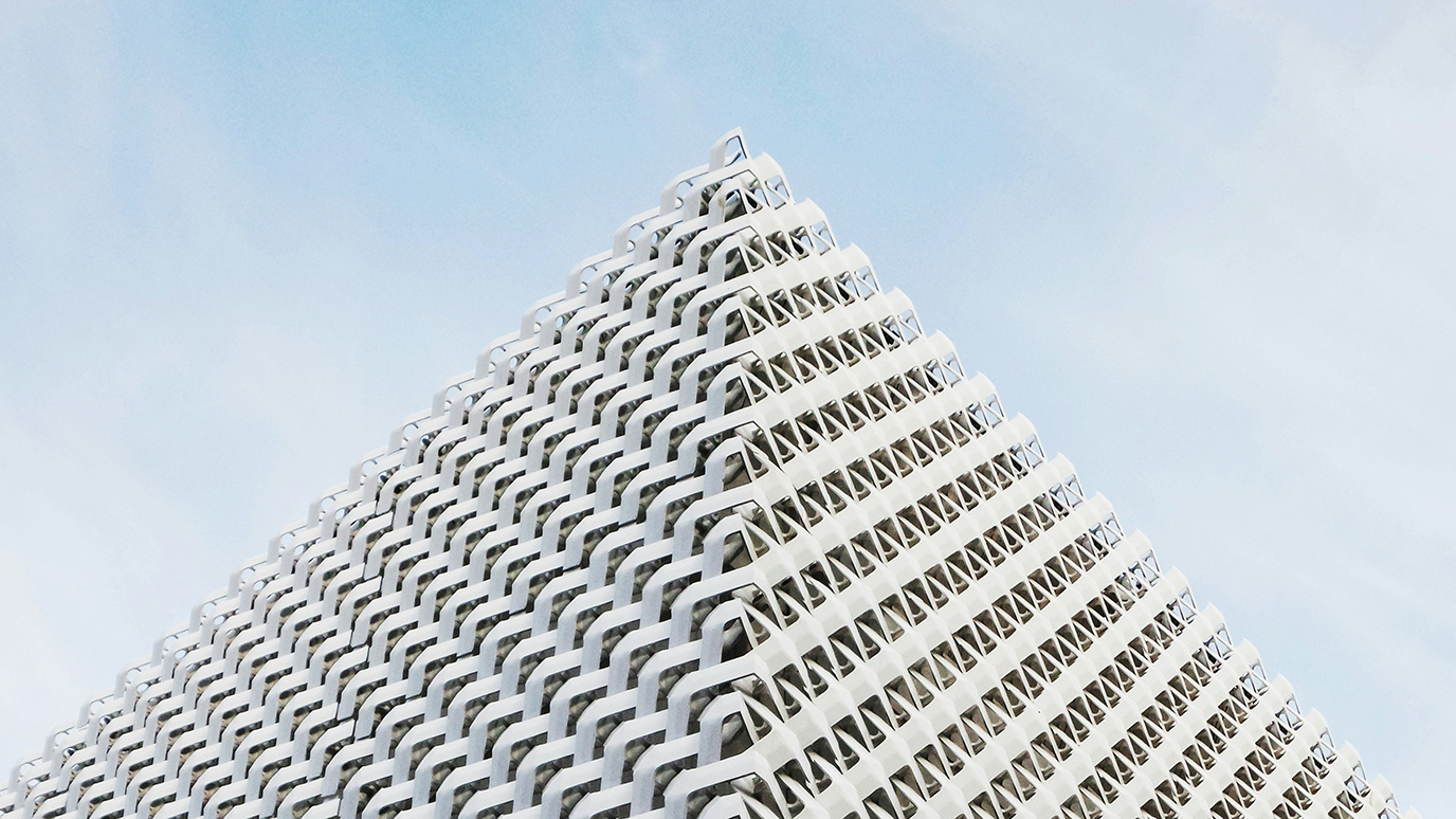 A tall building with a textured building skin made of white metal, with a triangular pattern and a perspective angle showing the right side in a closeup shot, against a light blue sky.