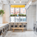 Modern laundromat with natural lighting, big windows, white tile floors, contemporary washers and dryers, tiled walls, bright accent wall, pendant light, and exposed ceilings.