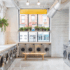 Modern laundromat with natural lighting, big windows, white tile floors, contemporary washers and dryers, tiled walls, bright accent wall, pendant light, and exposed ceilings.