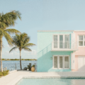 Key West design of a residential apartment building. Pastel blue and pink paint on the exterior of the building surrounded by the ocean in the background and in the foreground is a pool surrounded by lounge chairs and palm trees.