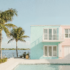 Key West design of a residential apartment building. Pastel blue and pink paint on the exterior of the building surrounded by the ocean in the background and in the foreground is a pool surrounded by lounge chairs and palm trees.