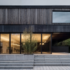 Modern house, wooden cladding combined with black metal accents and glass windows the entrance is framed in dark grey concrete stairs leading to an open air terrace with dining area, surrounded by greenery