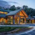 A modern, senior living facility with an exterior that features large windows and wooden accents, nestled in the woods. The entrance is decorated with beautiful flowers and plants, creating a welcoming atmosphere as you enter through its grand entryway and open parking lot.