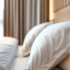 A close up photograph of a hotel bed slightly out of focusing highlighting the white and tan antimicrobial textiles. A large window covered with sheer curtains makes the room feel bright and airy.