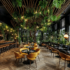 An image of biophilic restaurant design with wood floors, greenery in the ceiling panels, live walls and plants surrounding the edges of the restaurant. The bar on the left is trimmed in black and gold and round tables run parallel to the bar with a row of bench tables running along the right hand side of the room.